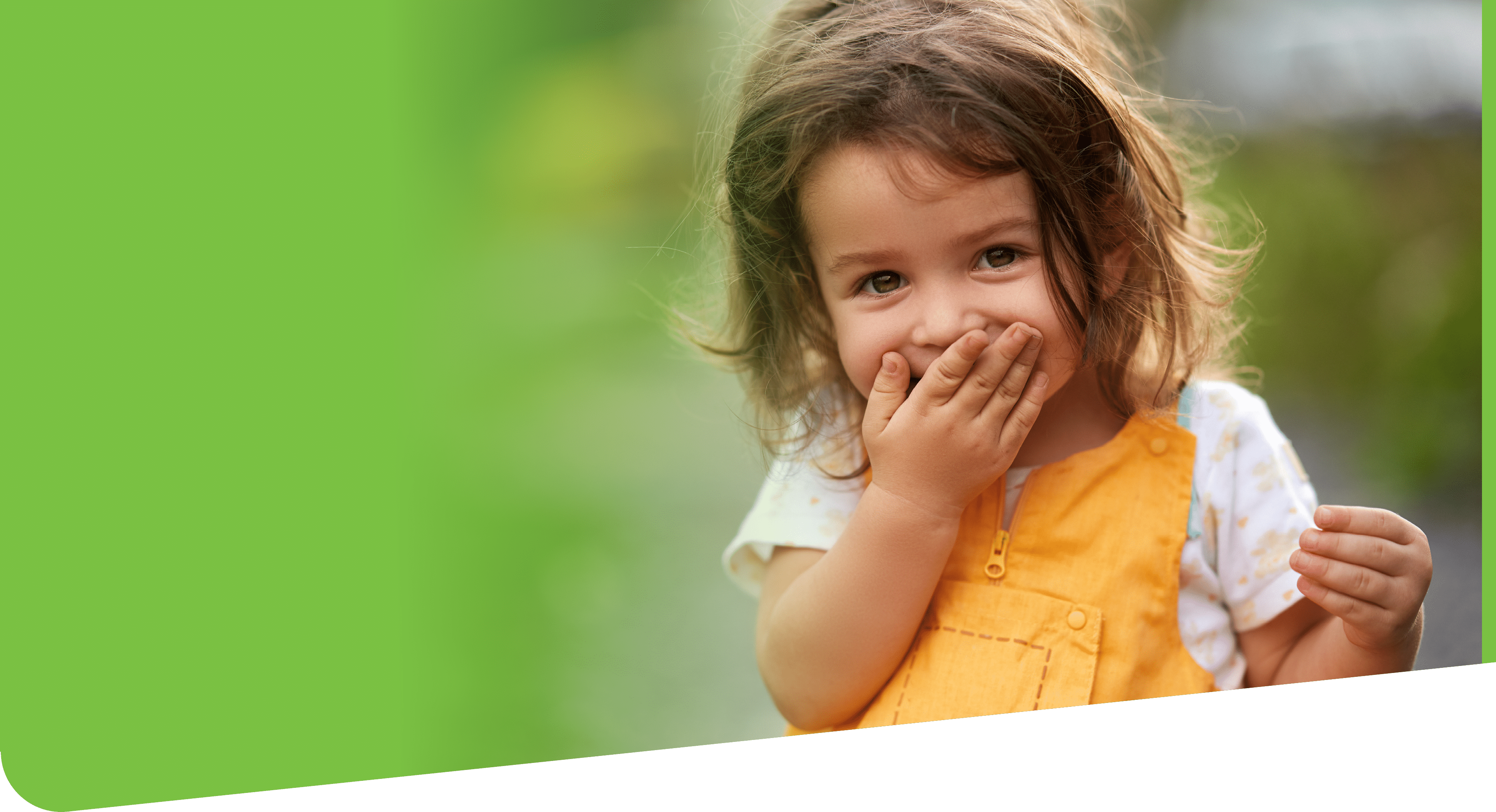 Little girl with brown hair outdoors, covering her mouth laughing