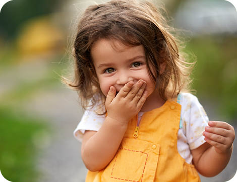 Little girl with brown hair outdoors, covering her mouth laughing