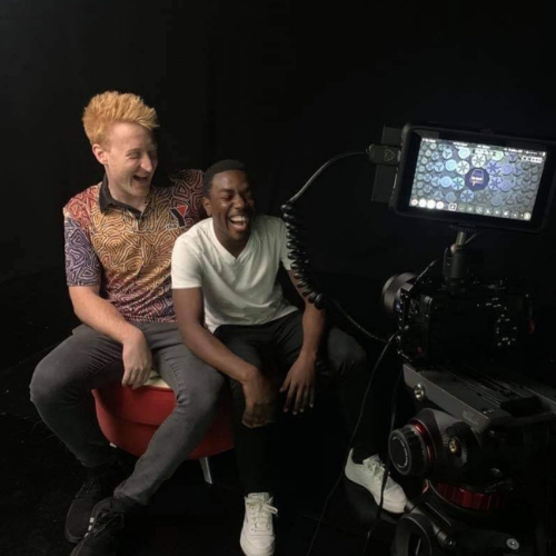Image of two people laughing, seated in front of a video camera