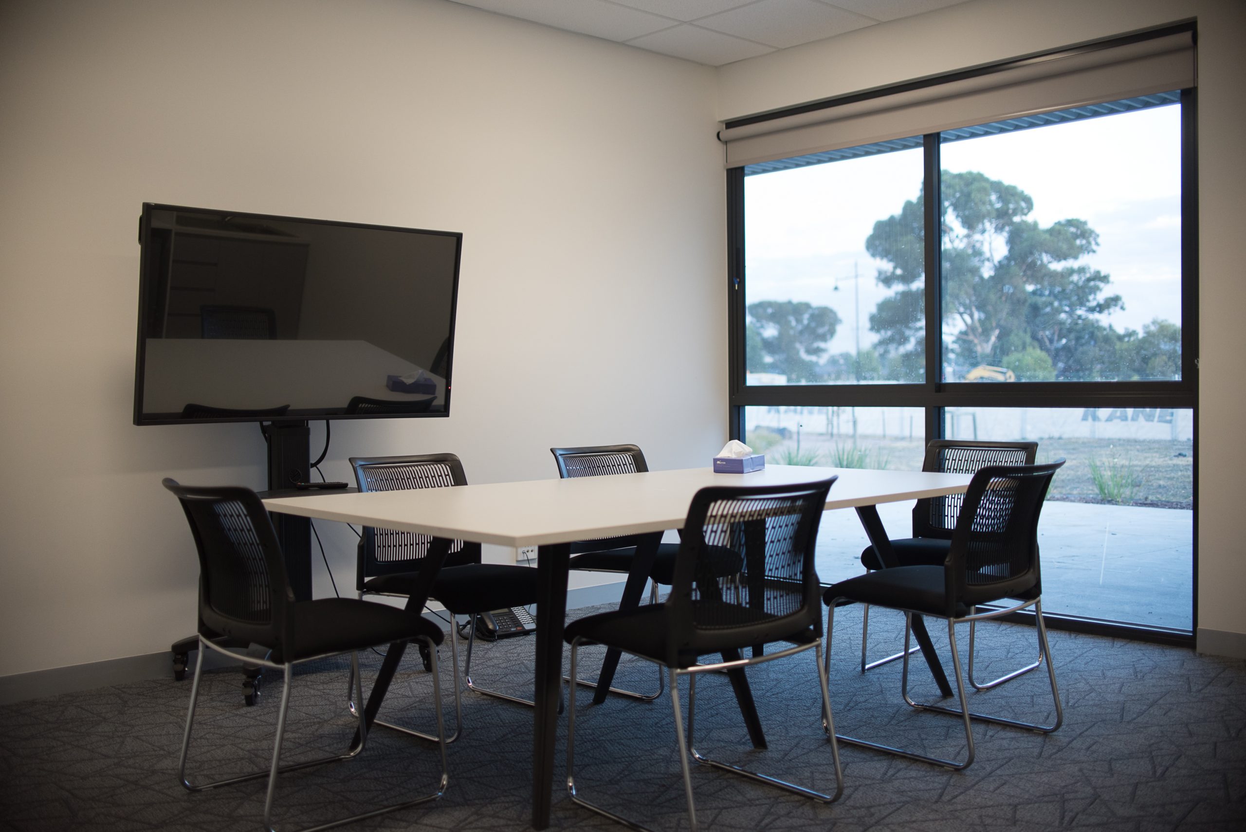 A clean carpeted meeting room with white table and 6 black chairs and a large monitor. Windows, with roller blinds, at one end look onto a courtyard.
