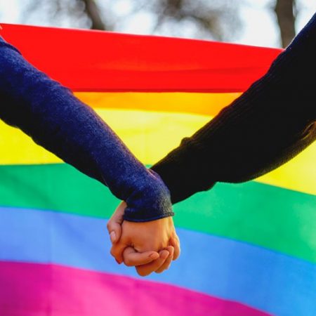 Image of the pride rainbow flag with two people holding hands in front of it (people not shown)